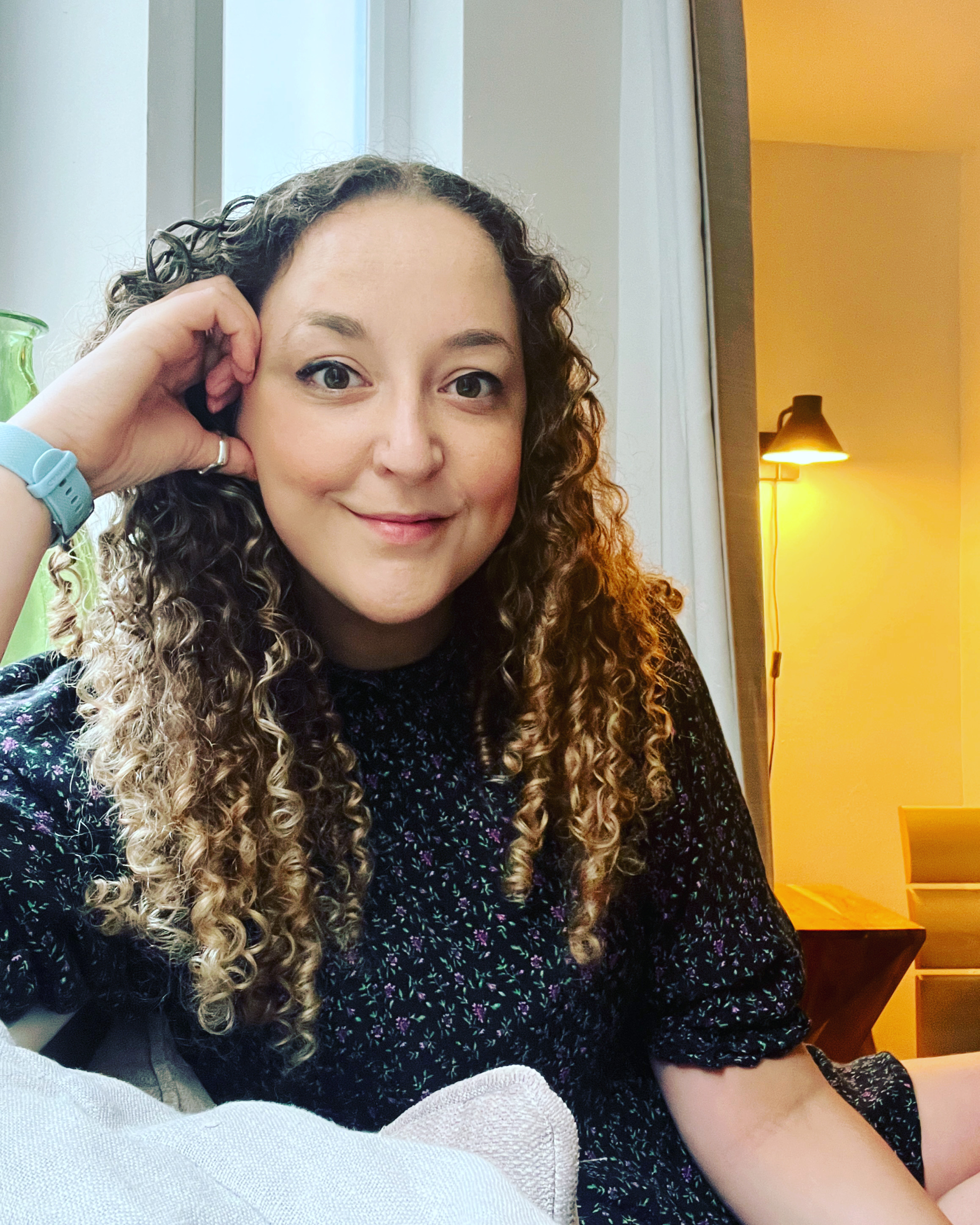 Hannah has curly hair, sitting indoors and smiling at the camera, resting her head on her hand. She is wearing a floral dress and is surrounded by a cozy, well-lit environment.